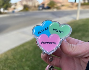 May include: A sparkly heart-shaped badge holder with three hearts. The hearts are blue, green, and pink. The blue heart says "Ativan", the green heart says "Versed", and the pink heart says "Propofol".