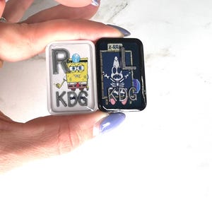 May include: Two rectangular pins featuring SpongeBob SquarePants and Patrick Star. One pin shows SpongeBob with the letter "R" and the letters "KDG". The other pin shows Patrick as an X-ray with the letters "KDG" and the text "X-RAY".