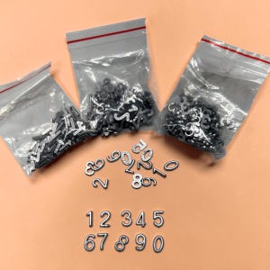 Peut inclure: Charmes de chiffres en métal argenté dans des sacs en plastique transparents et en vrac. Les chiffres de 1 à 0 sont affichés. Le fond est de couleur orange clair. Les breloques sont petites et adaptées à la fabrication de bijoux ou à l'artisanat.