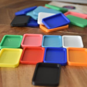 May include: A set of ten colorful square-shaped trays. The trays are made of plastic and are arranged in a pattern on a wooden surface. The colors of the trays are yellow, red, blue, green, orange, black, pink, white, light blue, and dark blue.