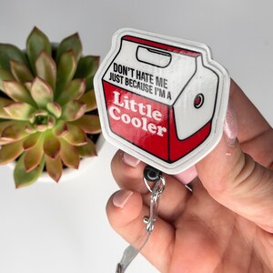 May include: A white and red badge reel with the text "DON'T HATE ME JUST BECAUSE I'M A Little Cooler". The badge reel is in the shape of a cooler. The badge reel is attached to a clear plastic strap.