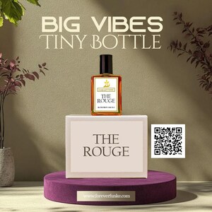 Puede incluir: Un peque&ntilde;o frasco de perfume rectangular con tapa negra, etiquetado como "THE ROUGE". Se encuentra sobre una caja blanca con el mismo texto, colocada sobre un pedestal de terciopelo p&uacute;rpura. La imagen incluye el texto "BIG VIBES TINY BOTTLE".
