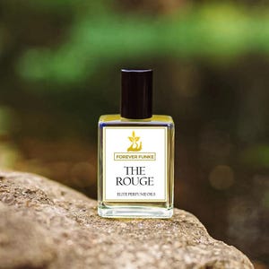 Puede incluir: Un frasco de perfume de vidrio transparente con una tapa negra, etiquetado "THE ROUGE" por Forever Funke. La etiqueta blanca presenta texto dorado y un logotipo dorado. La botella se encuentra sobre una superficie de piedra marr&oacute;n rugosa contra un fondo verde borroso.