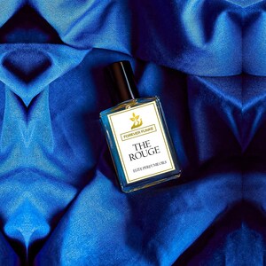 Puede incluir: Un frasco de perfume rectangular con tapa negra y etiqueta blanca con la inscripci&oacute;n "FOREVER FUNKE" y "THE ROUGE ELITE PERFUME OILS". El frasco contiene un l&iacute;quido claro y descansa sobre una tela de sat&eacute;n azul.