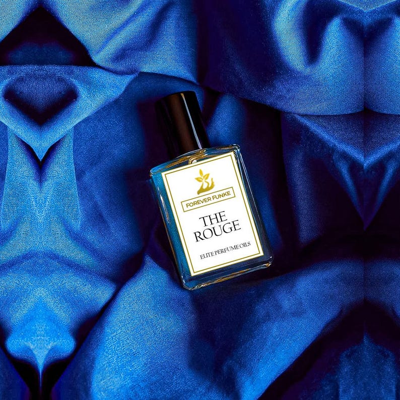 Puede incluir: Un frasco de perfume rectangular con tapa negra y etiqueta blanca con la inscripci&oacute;n "FOREVER FUNKE" y "THE ROUGE ELITE PERFUME OILS". El frasco contiene un l&iacute;quido claro y descansa sobre una tela de sat&eacute;n azul.