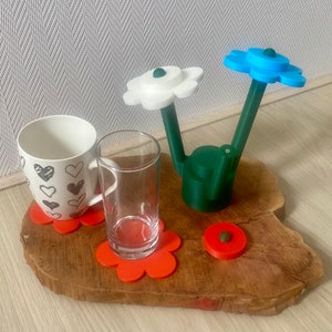 May include: A wooden tray with three red flower-shaped coasters. A white mug with black hearts, a clear glass, and a green teapot with blue and white flower-shaped lids are on the coasters.