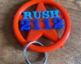 Könnte beinhalten: Ein roter, 3D-gedruckter Schlüsselanhänger mit einer Sternform und einem Kreis darum. Der Text "RUSH 2112" ist in Blau und Hellblau auf den Stern gedruckt.