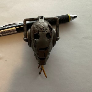 May include: A weathered gray and silver robot head with yellow wires. The robot has a large, round head with a face that is cracked and chipped. The robot's eyes are black and round. The robot's mouth is open in a slight smile.