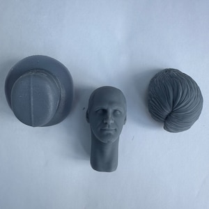 May include: Three gray resin model pieces: a hat, a bald head, and a sculpted hair piece. The head has detailed facial features. The hat is a rounded shape with a brim. The hair piece is a sculpted, wavy style.
