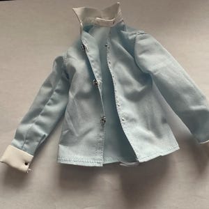 May include: A light blue button-down shirt with a white collar and cuffs. The shirt has long sleeves and decorative buttons. The garment is made of a soft fabric and is designed for a doll.