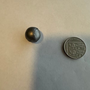 Könnte beinhalten: Ein kleiner, metallisch grauer Ball mit Naht, neben einer britischen Zehn-Pence-Münze. Die Münze zeigt ein detailliertes Design und Text. Der Ball scheint aus einem dichten Material zu bestehen.