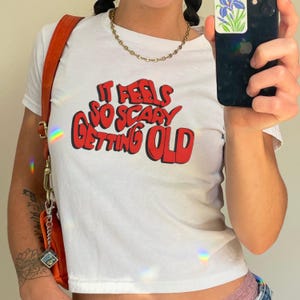 May include: White t-shirt with the text "IT FEELS SO SCARY GETTING OLD" in red, outlined in black. The shirt is short-sleeved and appears to be a crop top. An orange purse is visible on the left side of the image.