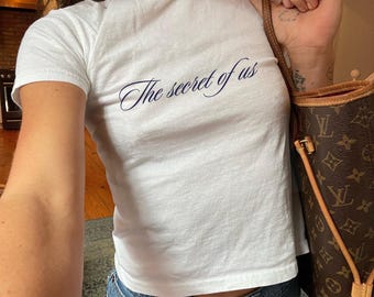 May include: White t-shirt with the text "The secret of us" in blue cursive lettering. The shirt is being worn by a person and is cropped at the waist.