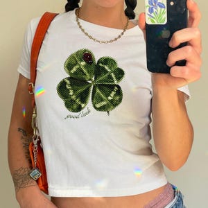 Puede incluir: Top corto blanco con un gran gráfico de trébol de cuatro hojas verde, una mariquita y la frase "good luck". La camiseta tiene mangas cortas y cuello redondo. También se ven un collar de cadena dorada y un bolso naranja.
