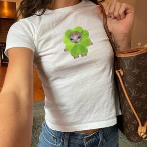 May include: White crop top t-shirt featuring a graphic of a kitten inside a green four-leaf clover. The kitten has a light brown and white fur. The t-shirt is made of a soft, lightweight material.