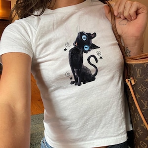 Könnte beinhalten: Weißes T-Shirt mit einem Cartoon-Design einer schwarzen Katze mit großen blauen Augen, umgeben von Sternen und Wirbeln. Die Katze sitzt mit eingerolltem Schwanz. Das T-Shirt hat kurze Ärmel und einen Rundhalsausschnitt.