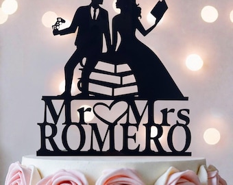 Op de afbeelding: Een zwarte taarttopper met een silhouet van een persoon in een pak met een gamecontroller en een persoon in een jurk met een boek, beide zittend op een stapel boeken. De tekst luidt Mr & Mrs ROMERO met een hartvorm in het midden.