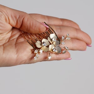 A hand holding a gold-colored hair comb with a floral design and small gemstones.