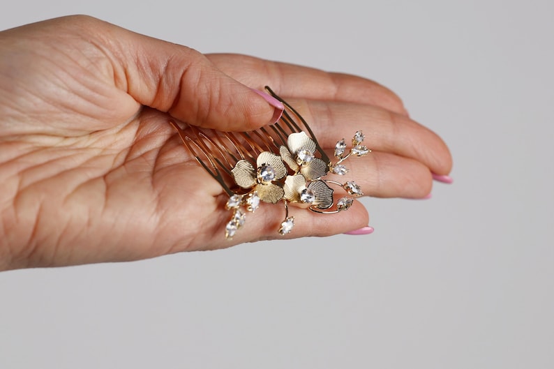 A hand holding a gold-colored hair comb with a floral design and small gemstones.