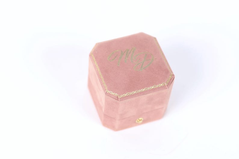 A pink velvet ring box with the initials 'M.D.' embossed on it.