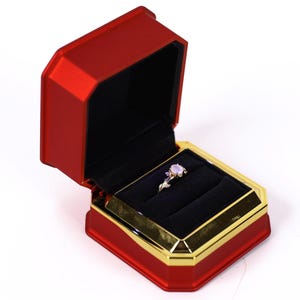 A red and gold jewelry box containing a ring, placed on a white background.