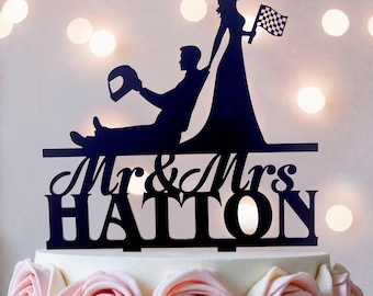Könnte beinhalten: Ein schwarzer Tortenstecker mit der Silhouette einer sitzenden Person, die einen Rennhelm hält, und einer stehenden Person mit einer Zielflagge. Unter den Figuren steht der Text Mr & Mrs HATTON in einer Kombination aus Schreib- und Blockschrift.