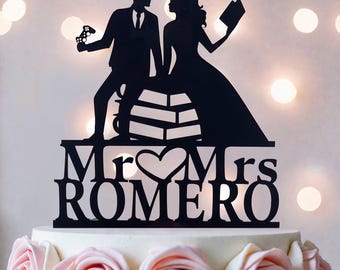 May include: Black acrylic wedding cake topper featuring a bride and groom silhouette. The groom holds a game controller, the bride holds a book. The topper reads "Mr & Mrs ROMERO" with a heart symbol. The cake is decorated with pink roses.