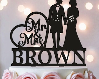 May include: A black cake topper featuring a silhouette of a couple, with the person on the left wearing a kilt and the person on the right in a gown. A heart shape contains the text Mr & Mrs, and the base displays the name BROWN.