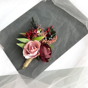 May include: A small bouquet of artificial flowers in shades of pink and burgundy. The bouquet includes roses, berries, and greenery. It is tied with twine and would be suitable for a wedding or other special occasion.