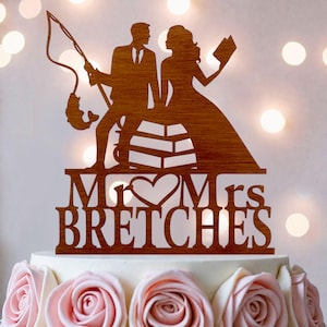 Puede incluir: Un adorno de madera marrón para pastel con la silueta de una persona pescando y otra sosteniendo un libro, sentadas sobre una estructura. El texto dice Mr & Mrs BRETCHES con un corazón entre las palabras, todo en un diseño recortado.