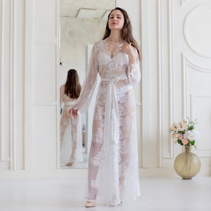 May include: A white lace robe with long sleeves and a sash. The robe is sheer and has a delicate floral pattern. The model is standing in a white room with a mirror in the background.