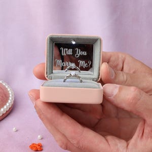 May include: A light pink ring box with a gray interior lining and a small LED light. The inside lid features a mirror with the engraved text Will You Marry Me? A silver-toned ring with a central stone is positioned in the center slot of the box.