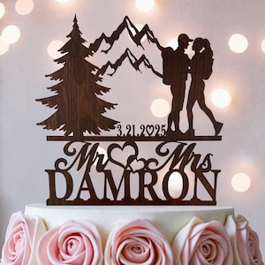 May include: A dark brown wooden cake topper featuring a silhouette of a pine tree, mountain range, and two hikers with backpacks. The text 3.21.2025, Mr & Mrs, and the name DAMRON are laser-cut into the wood, accented by two heart shapes.