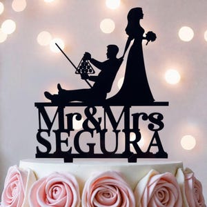 May include: A black cake topper featuring a silhouette of a person sitting and holding a pool cue with a rack of billiard balls, and a standing person holding a bouquet. The text Mr & Mrs SEGURA is displayed in a bold font at the base.