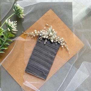 May include: A silver and white bridal hair comb with a large crystal and pearl accents. The comb is on a brown and gray background.