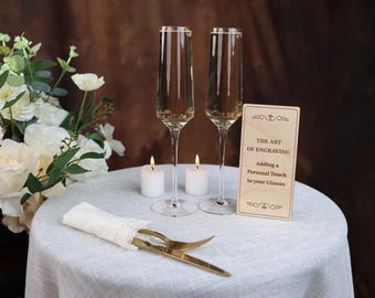 May include: Two champagne flutes with a wooden sign that reads "The Art of Engraving, Adding a Personal Touch to your Glasses". The flutes are filled with a golden liquid and are sitting on a white tablecloth with two white candles.