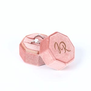 A pink velvet box containing a diamond ring, with the letters "JR" visible on the side of the box.
