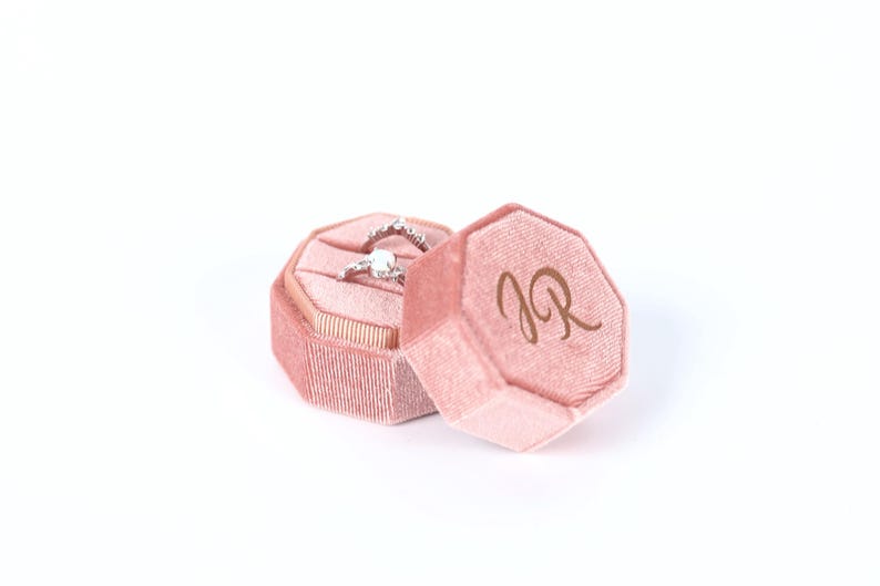 A pink velvet box containing a diamond ring, with the letters "JR" visible on the side of the box.