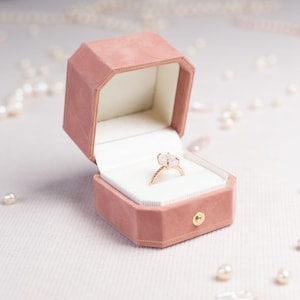 May include: An octagonal ring box in a dusty rose velvet finish with a gold-tone metal clasp, containing a gold ring with a rectangular white stone set in a four-prong setting. The interior of the box is lined with white fabric.