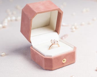May include: An octagonal ring box in a dusty rose velvet finish with a gold-tone metal clasp, containing a gold ring with a rectangular white stone set in a four-prong setting. The interior of the box is lined with white fabric.