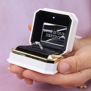 Könnte beinhalten: Eine weiß-goldene Ringbox mit schwarzem Innenraum, ausgestattet mit einem kleinen LED-Licht und einem Spiegel mit der Gravur LOVE IS OUR DESTINY. Ein silberner Ring mit einem klaren Stein befindet sich in der Mitte des dunklen Samtschlitzes.