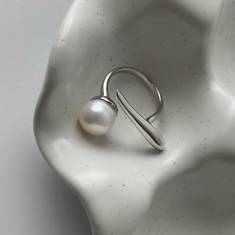 derMomentø - Handcrafted large pearl open ring with a naturally grown freshwater baroque pearl, set on a white rhodium-plated silver band. Display on a ceramic tray on a clean background.