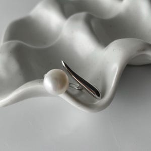derMomentø - Handcrafted large pearl open ring with a naturally grown freshwater baroque pearl, set on a white rhodium-plated silver band. Display on a ceramic tray on a clean background.