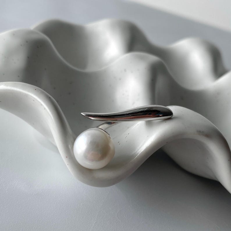 derMomentø - Handcrafted large pearl open ring with a naturally grown freshwater baroque pearl, set on a white rhodium-plated silver band. Display on a ceramic tray on a clean background.
