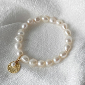 Könnte beinhalten: Ein Armband mit cremeweißen, unregelmäßig geformten Perlen. Ein kleiner, strukturierter goldfarbener Anhänger mit einer kleineren Perle hängt am Armband. Das Armband wird auf einer weißen, strukturierten Oberfläche präsentiert.