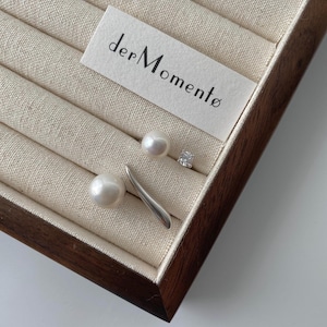 May include: A wooden box with a white label that says "der Momente" and three pairs of earrings. The earrings are made of silver and pearls.
