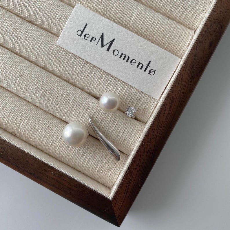May include: A wooden box with a white label that says "der Momente" and three pairs of earrings. The earrings are made of silver and pearls.