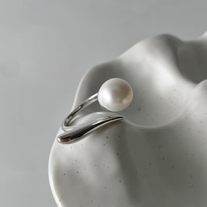 derMomentø - Handcrafted large pearl open ring with a naturally grown freshwater baroque pearl, set on a white rhodium-plated silver band. Display on a ceramic tray on a clean background.