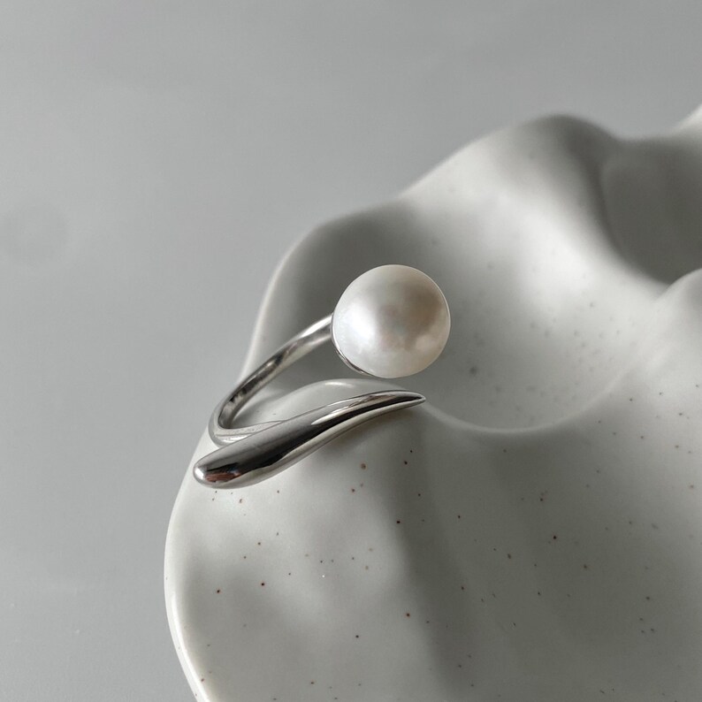 derMomentø - Handcrafted large pearl open ring with a naturally grown freshwater baroque pearl, set on a white rhodium-plated silver band. Display on a ceramic tray on a clean background.