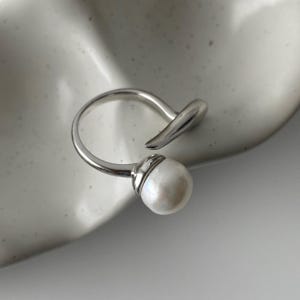 derMomentø - Handcrafted large pearl open ring with a naturally grown freshwater baroque pearl, set on a white rhodium-plated silver band. Display on a ceramic tray on a clean background.